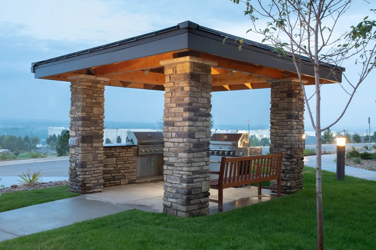 The image features an inviting outdoor area with sturdy stone pillars, a well-equipped grill station, and a comfortable wooden bench. The setting is designed for barbecuing, socializing, and relaxing, creating a welcoming environment where guests can gather and enjoy meals together. The scene exudes a warm, communal atmosphere perfect for unwinding after a day of exploring local attractions.