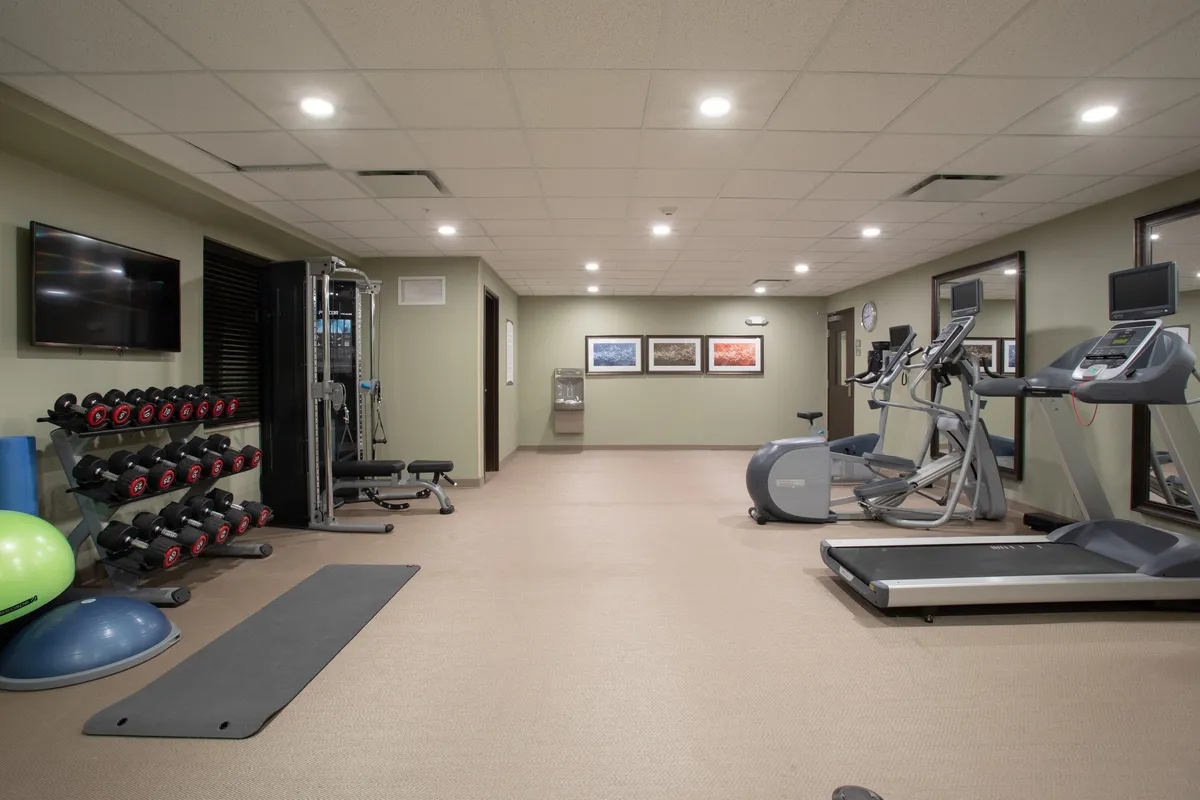 A bright, motivational fitness center equipped with a full dumbbell rack, treadmill, elliptical machine, and weight training equipment. Artwork adorns the wall, and there’s a water cooler nearby, suggesting both functionality and a pleasant workout environment.