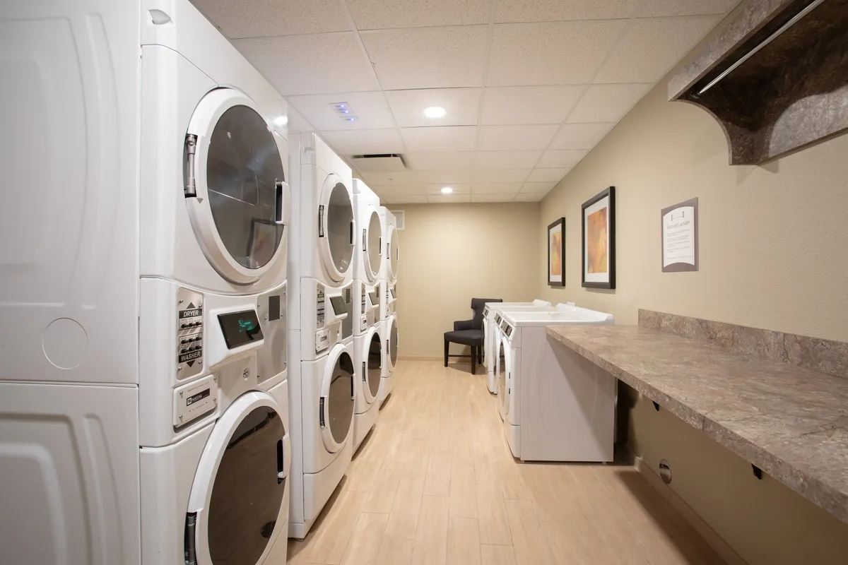 A practical yet comfortable laundry area inside the property, equipped with stacked washing machines, a convenient folding table, a cushioned chair, and decorative wall art. The space is clean, functional, and welcoming, enhancing longer or family stays.