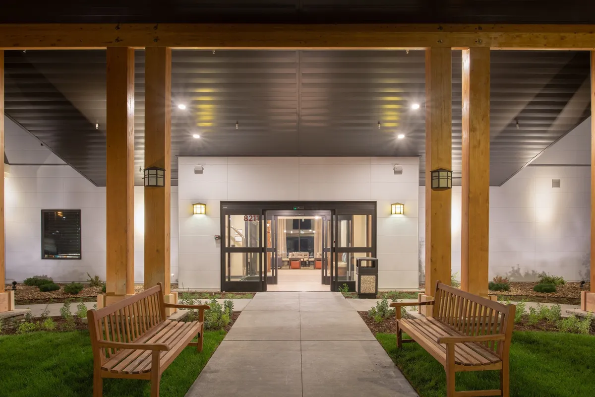 This exterior image showcases the entrance door of Staybridge Suites, flanked by wooden benches and surrounded by lush, landscaped greenery. Soft evening lighting creates an inviting atmosphere where guests can gather, relax, and enjoy tranquil moments outdoors.