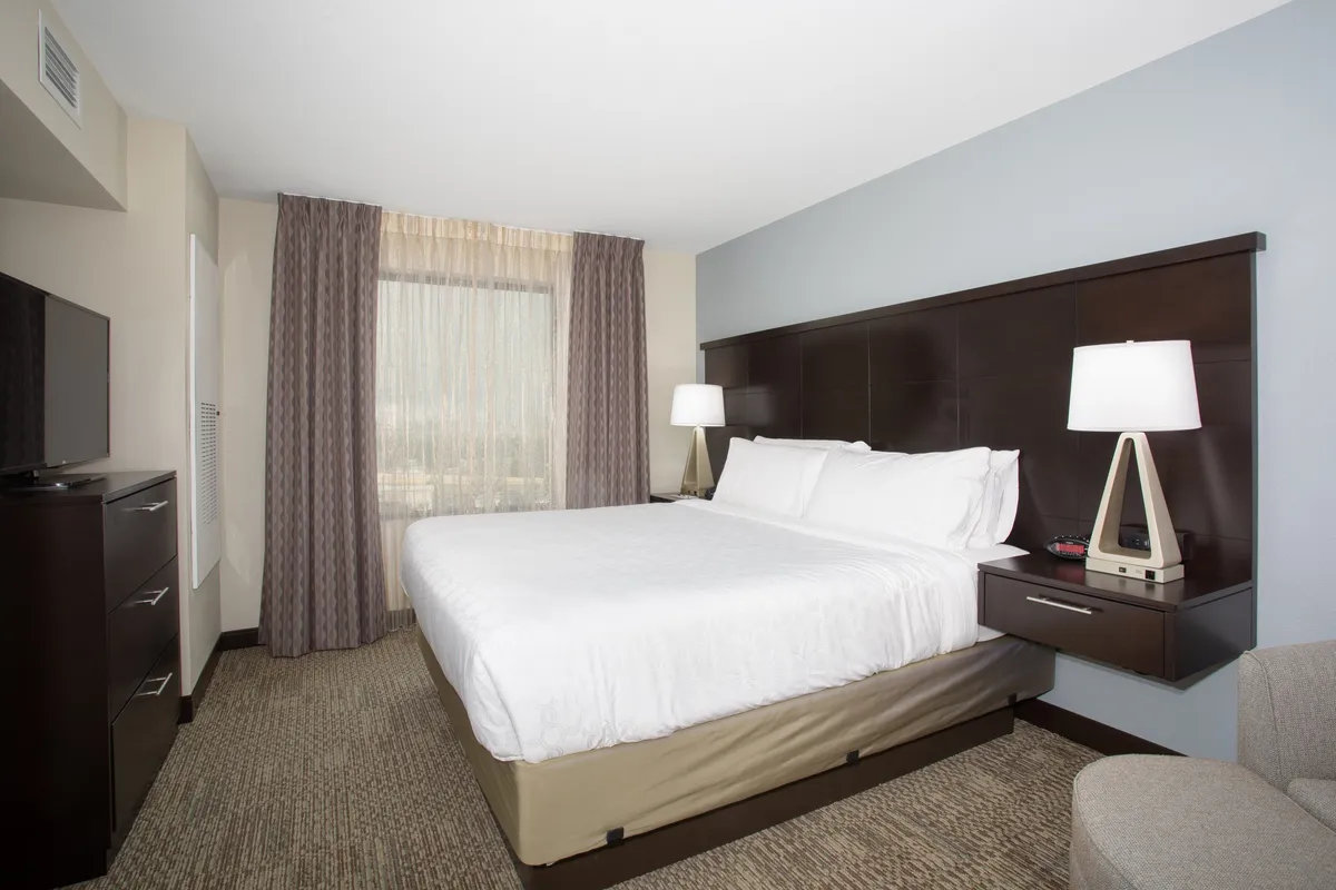 A bright bedroom featuring a plush queen bed, a large window letting in natural light, a modern nightstand, and a flat-screen television mounted on the wall. The decor is soothing, with soft textiles and uncluttered surfaces that evoke comfort and privacy.